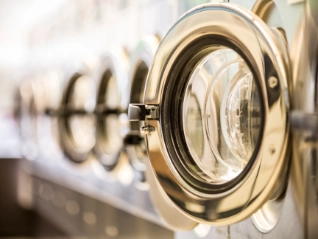  Close-up of a row of commercial washing machine doors in a laundromat, with the first chrome door open and the others receding in a blurred...