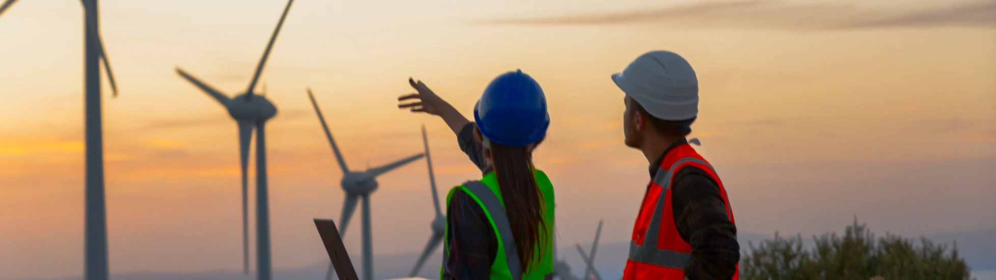 Two wind technicians (green vest/blue hard hat pointing with laptop; orange vest/white hard hat) overlook a field of large wind turbines at sunset.